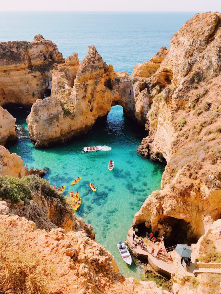 A bird’s eye view of a sandy yellow cove surrounding sparkling blue waters and people in kayaks in Lagos, Portugal.