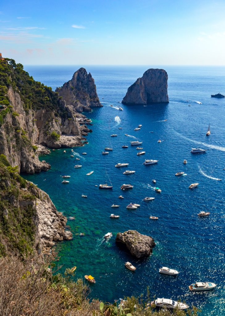 The view from a rugged Italian cliff edge of sparkling blue waters filled with white boats.
