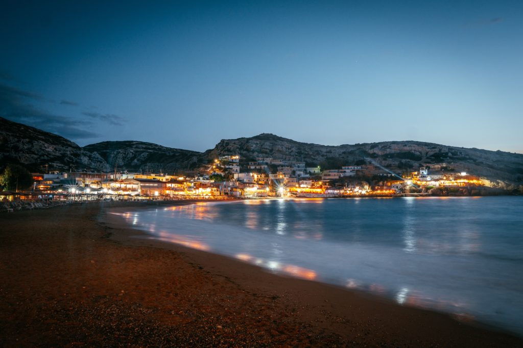 A view across the beach and still sea to a coastal Greek town all lit up at night.