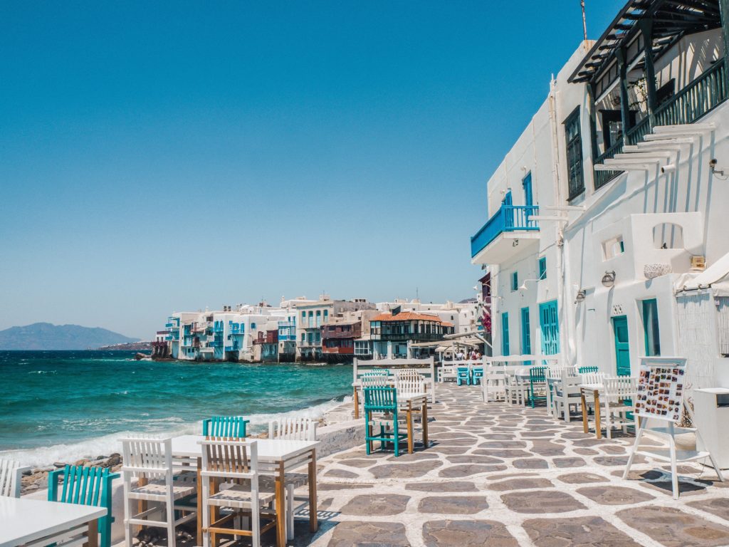 White and blue buildings line the Greek island seafront with simple tables and chairs for outdoor dining.