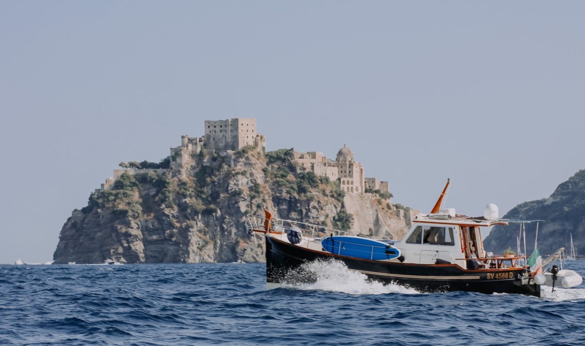 In the foreground a boat motors past, in the background a historical castle and fortress covers a small, rocky Italian island.
