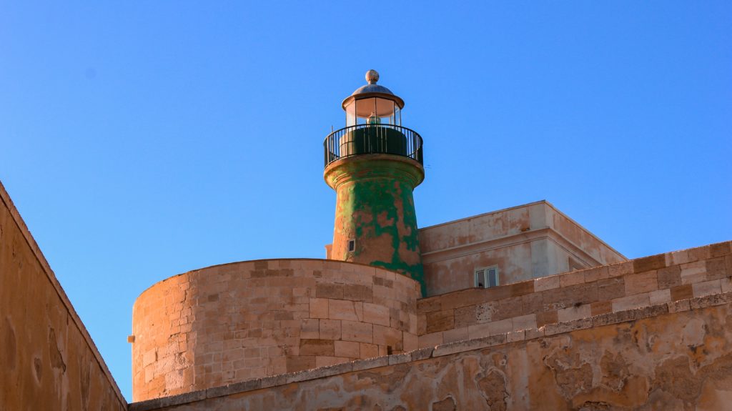 Against a clear blue sky is a historical watchtower with peeling green paint on the mediaeval walls of Syracuse in Sicily, one of the best Italian islands for history.